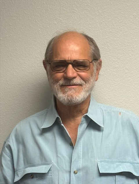 Smiling elderly man in light blue shirt with glasses and beard.