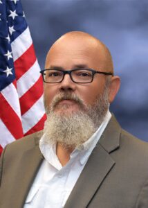 Portrait of a bald man with a beard in front of an American flag.