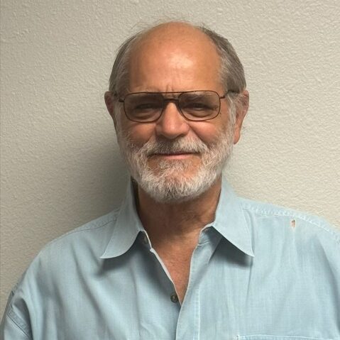 Smiling elderly man in light blue shirt with glasses and beard.